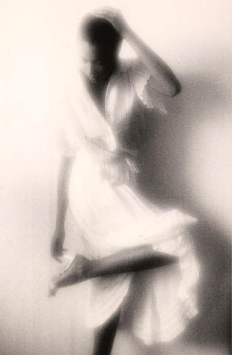 A woman in a light-colored dress poses with one leg raised and her hand on her head.