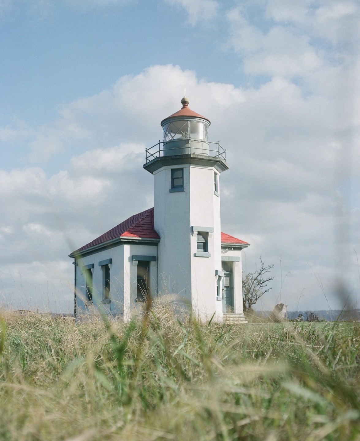 A white lighthouse with a red roof stands tall amidst a field of tall grass under a cloudy sky.