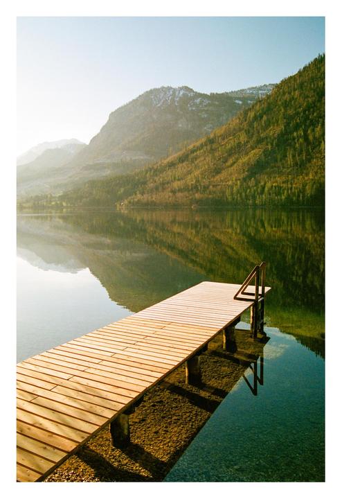 A wooden pier extends into a calm lake, reflecting the surrounding mountains and trees under a bright sky.