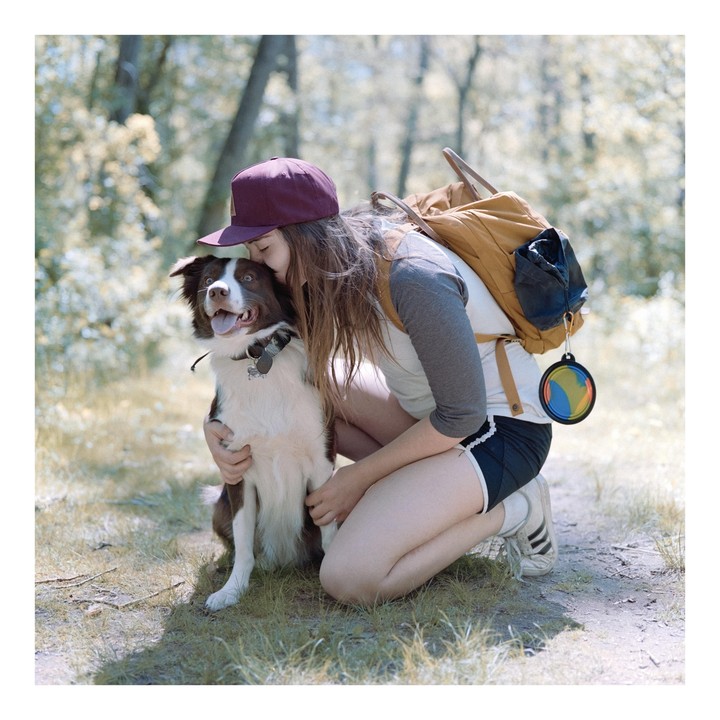 A woman wearing a backpack and a hat kneels to hug a dog on a dirt path in a forest.