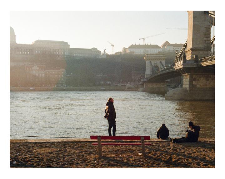 Three people are on a sandy bank next to a river with a bridge and buildings in the background.