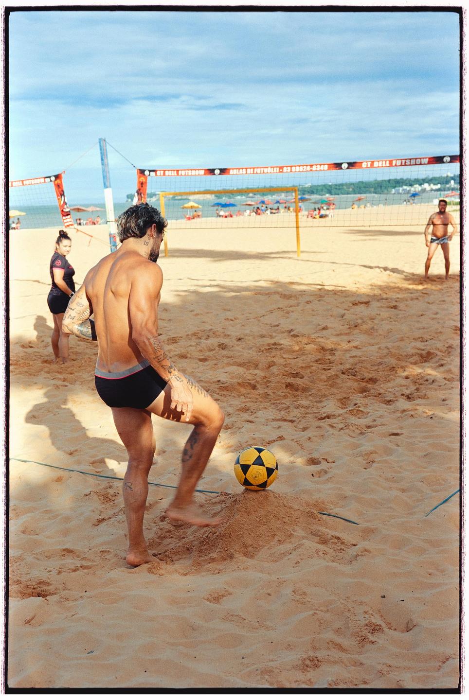 A man in swim trunks stands on a sandy beach with a yellow and black ball at his feet, with a volleyball net and other people in the background.