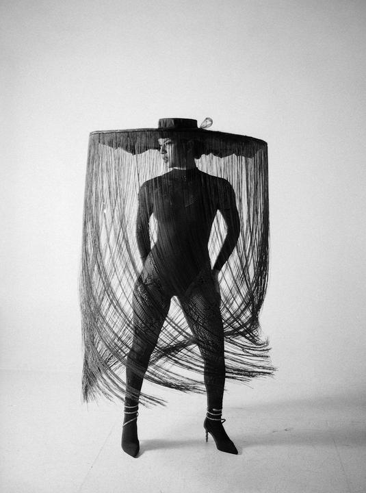 A woman in a full-body garment and a wide-brimmed hat with long fringe stands against a plain background.