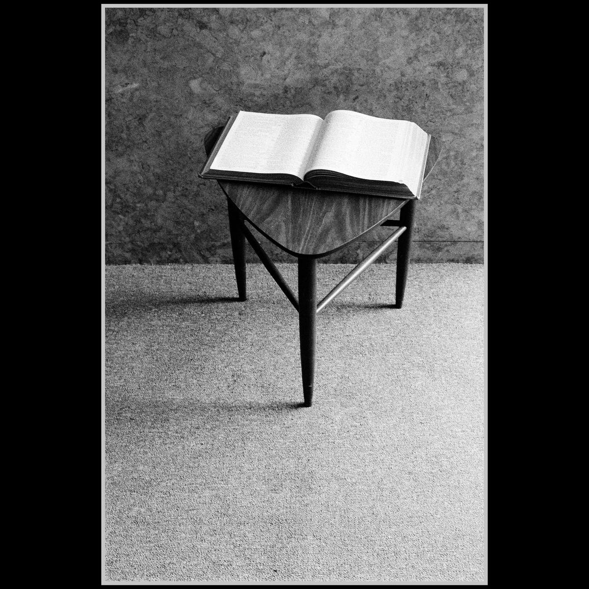 An open book rests on a small wooden table in a room with a textured wall and carpeted floor.