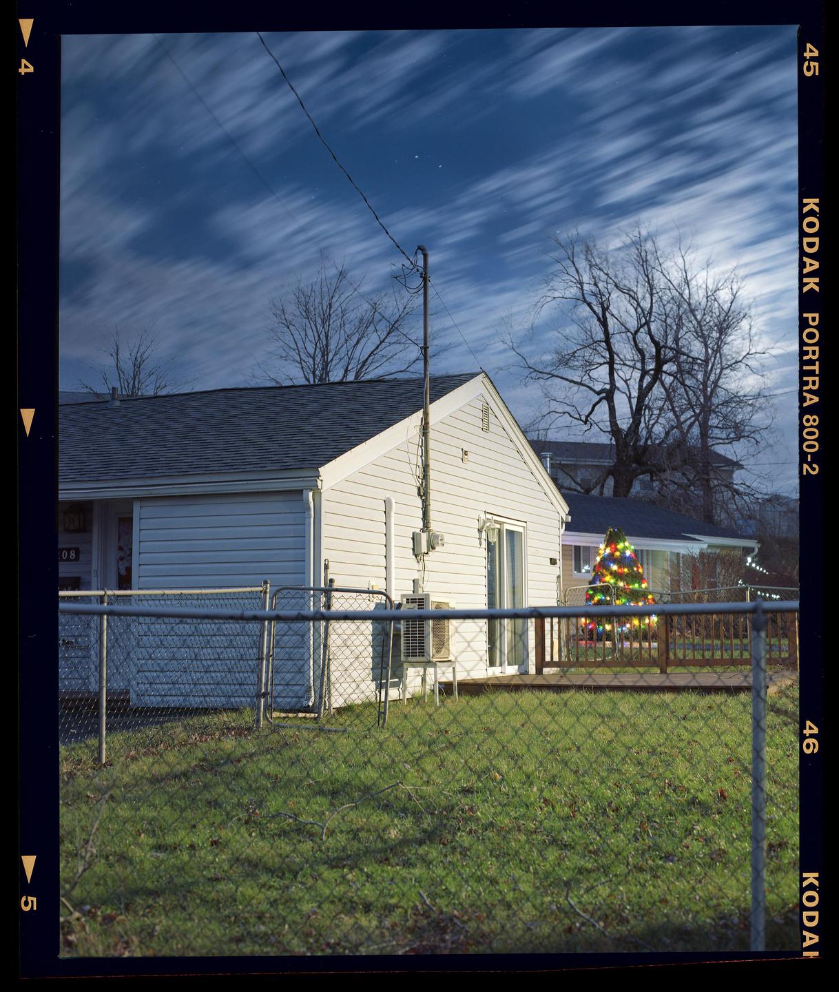 A white house with a chain link fence in the foreground is visible under a cloudy night sky, with a decorated Christmas tree in the background.