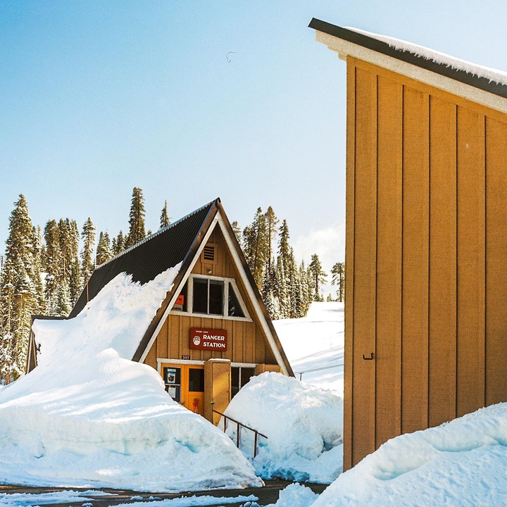 A ranger station with a triangular roof is surrounded by deep snow, with a forest of pine trees in the background.