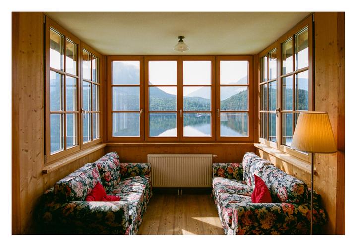 Two floral sofas with red pillows face each other in a wood-paneled room with large windows overlooking a lake and mountains.