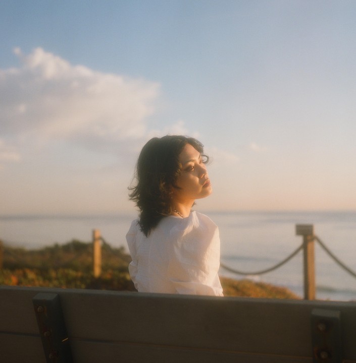 A woman sits on a bench overlooking the ocean at sunset.