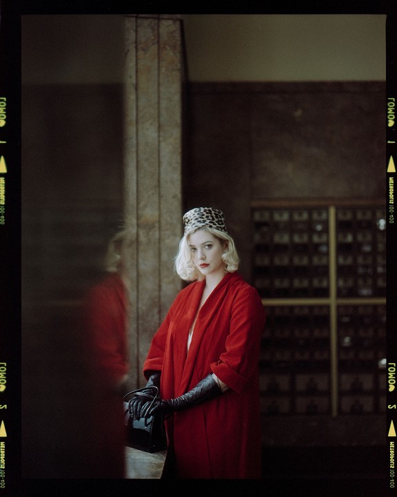 A woman in a red coat, black gloves, and a leopard print hat stands in a room with a reflection of herself visible on the left.
