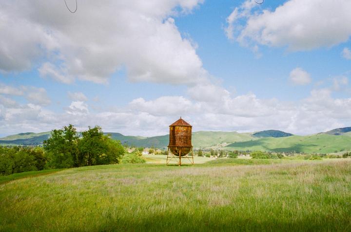 A rusty water tower stands in a grassy field with rolling hills and a blue sky with clouds in the background.