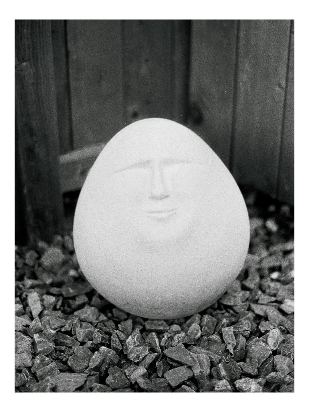 A round, smooth stone with a face carved into it rests on a bed of gravel.