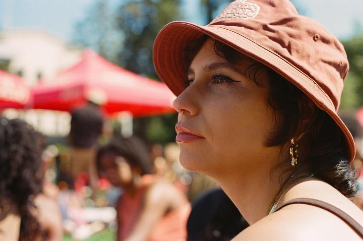 A woman in a bucket hat looks to the right with other people and a red tent in the background.
