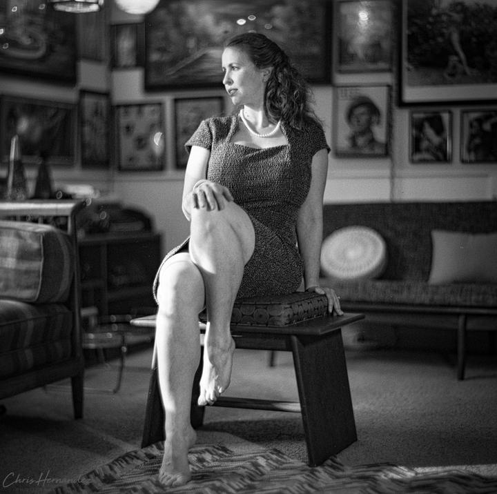 A woman wearing a dress and pearl necklace sits on a stool in a room with many framed pictures on the wall.