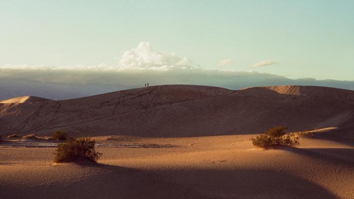 Two people stand on a distant sand dune in a desert landscape with scattered bushes and mountains under a cloudy sky.