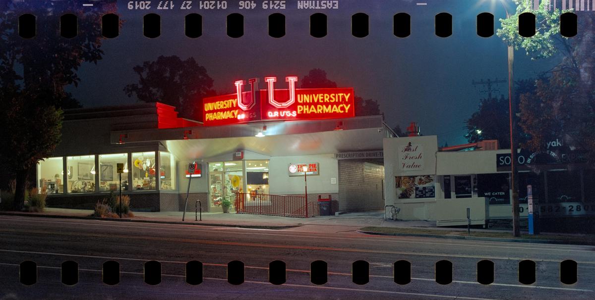 A pharmacy with a bright red neon sign is visible from across the street at night.