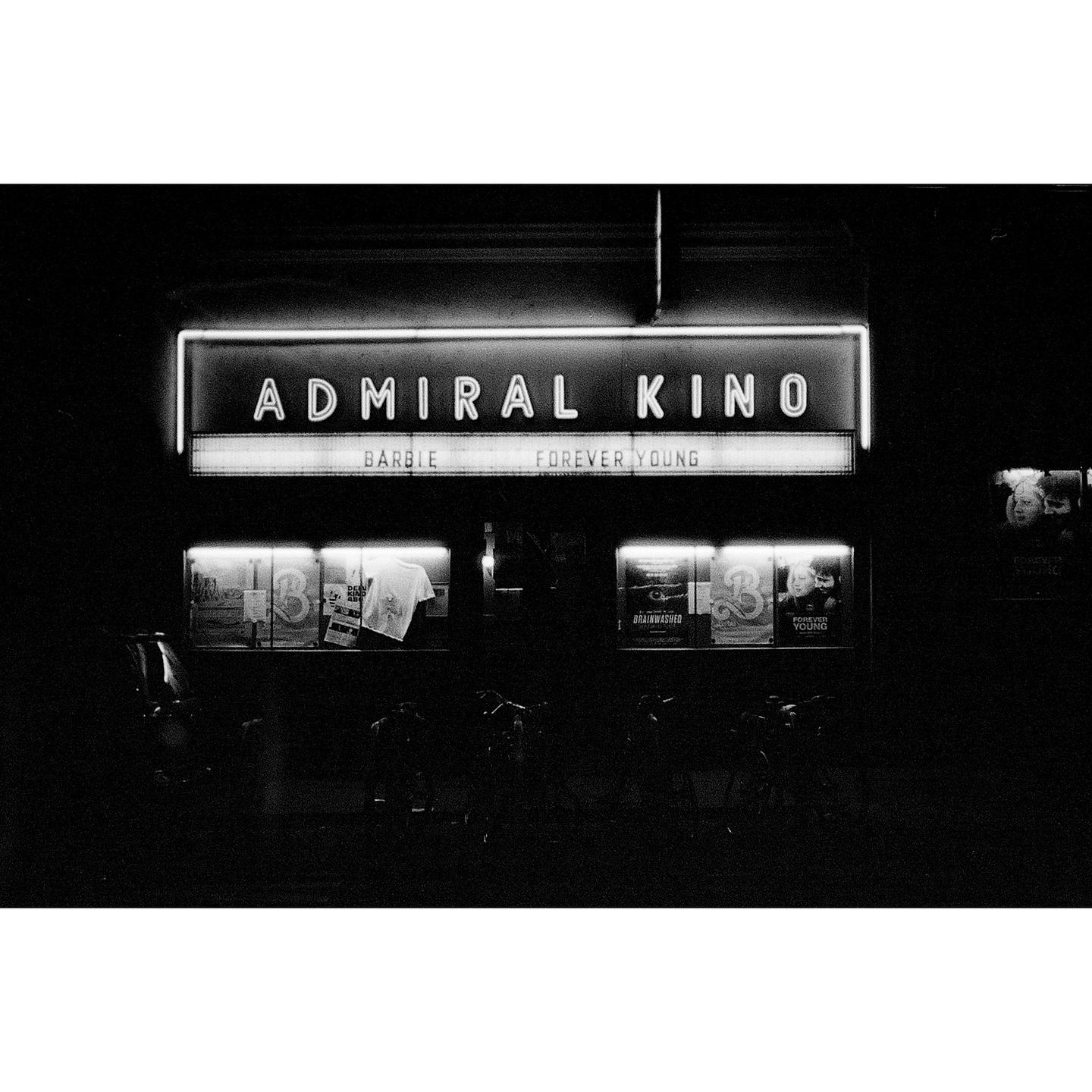 A neon sign for "Admiral Kino" is illuminated at night, with movie posters visible below.