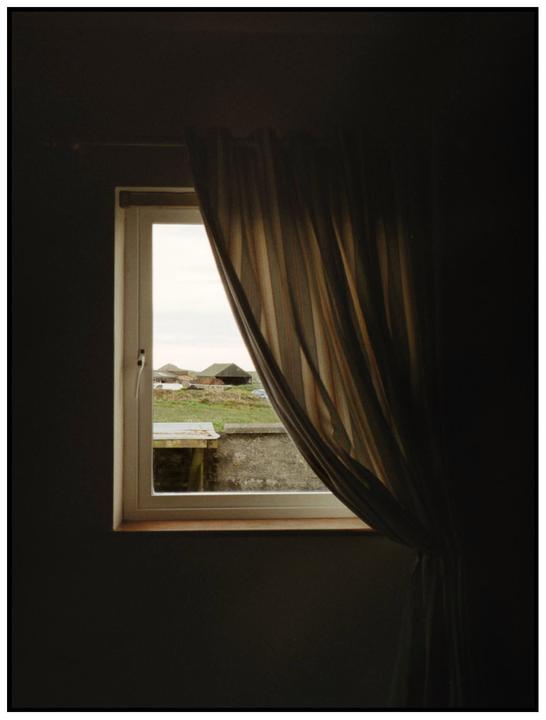 A window with a striped curtain pulled to one side reveals a view of a grassy field and buildings outside.