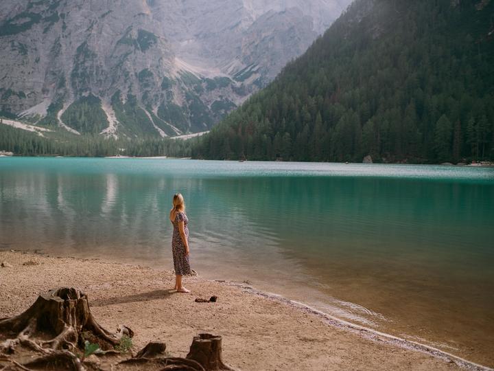 A woman stands barefoot on the shore of a turquoise lake, looking out at the water and mountains.
