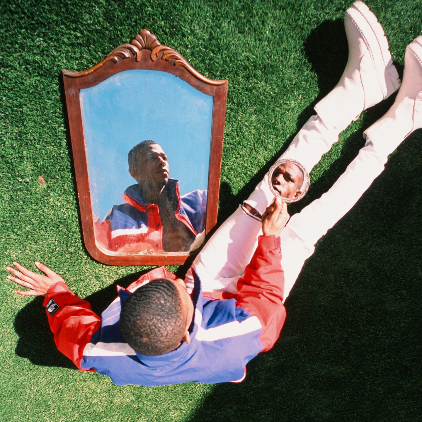 A man in a red, white, and blue jacket lies on green grass, looking at his reflection in a small handheld mirror while a larger ornate mirror reflects his upper body and the blue sky.