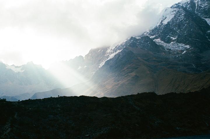 Sunlight breaks through clouds over a mountain range with snow-capped peaks and a dark ridge in the foreground.