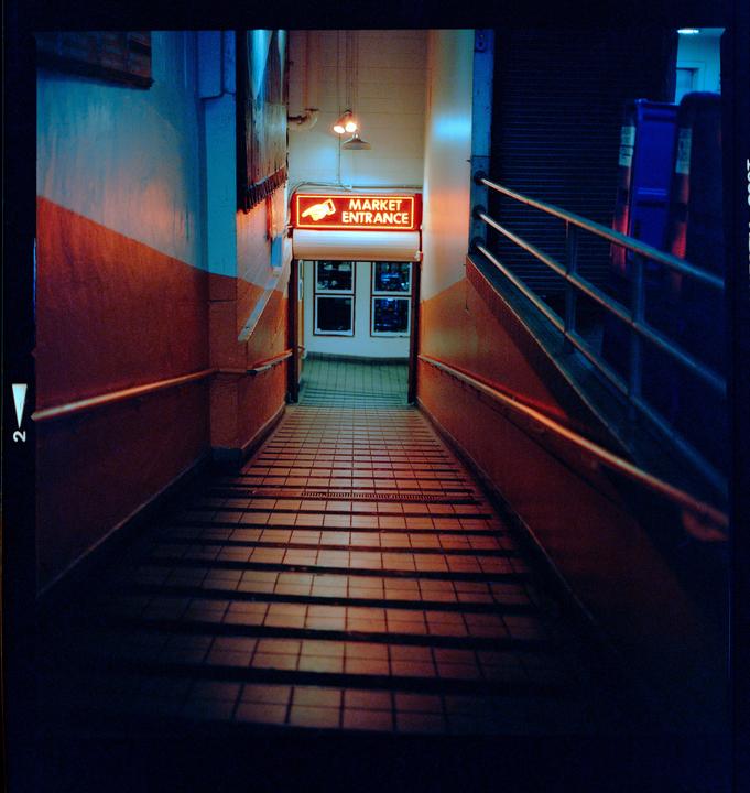 A tiled ramp leads down to a market entrance sign illuminated by neon light.