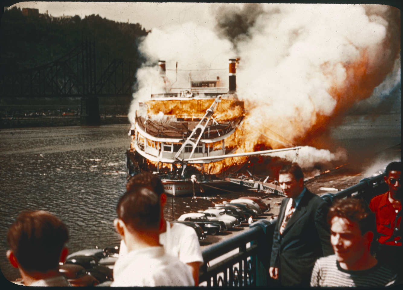 A steamboat is engulfed in flames and smoke on a river while people watch from a bridge.