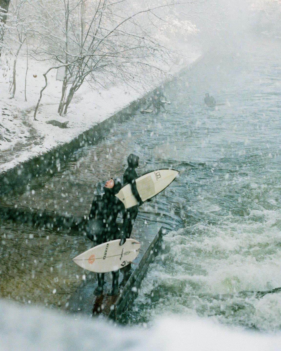 Two surfers in wetsuits stand on the bank of a river holding their surfboards while it snows.