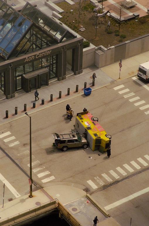 An overturned yellow truck and a car with an open trunk are on a street next to the Aon Center building.