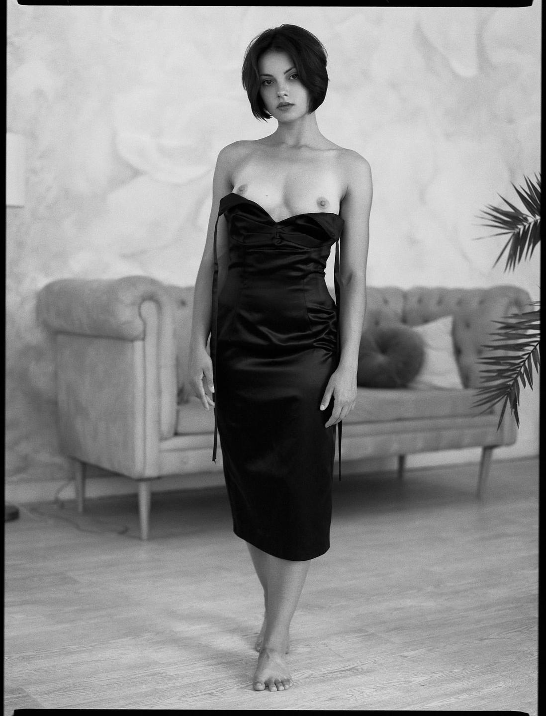 A woman in a black dress stands barefoot in a room with a sofa and a plant.