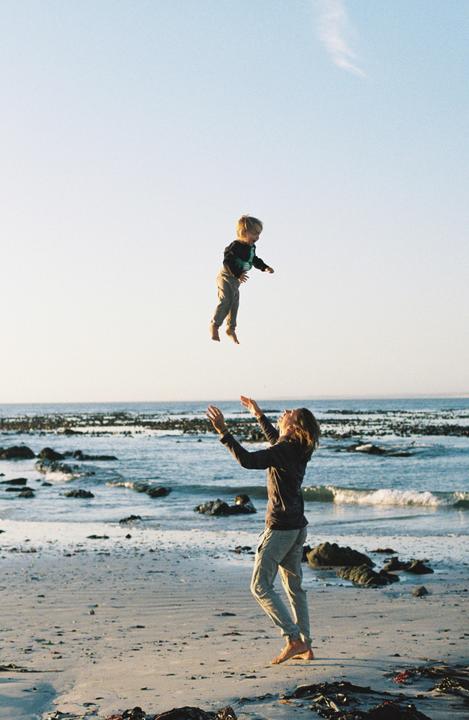 A man throws a child into the air on a rocky beach at sunset.