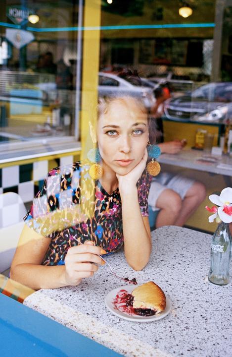A woman with large earrings sits at a diner table with a slice of pie, looking directly at the viewer through a window.