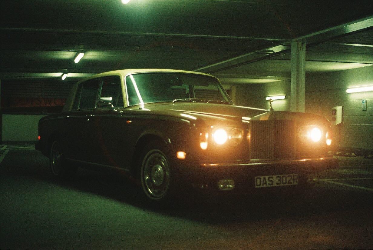 A classic car with its headlights on is parked in an underground garage.