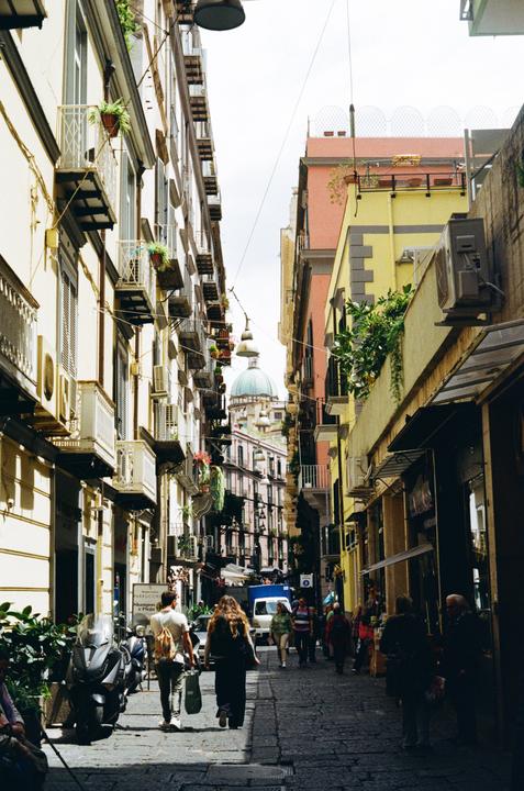 People walk down a narrow street lined with tall buildings and balconies, with a domed building visible in the distance.