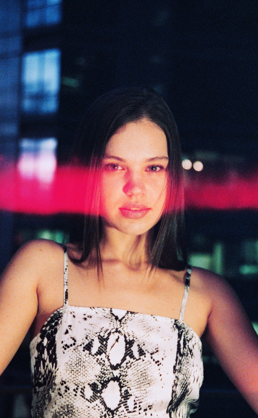 A woman with long dark hair and a snakeskin print top looks directly at the viewer, illuminated by a red light.