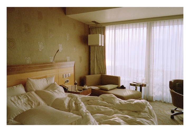 An unmade bed with white sheets and pillows sits in a hotel room with sheer curtains covering a large window.