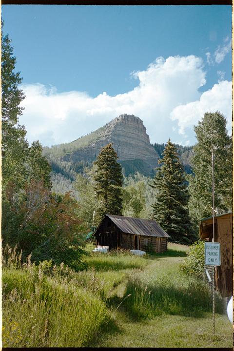 A small wooden cabin sits in a grassy field with a large mountain and trees in the background under a blue sky with clouds.