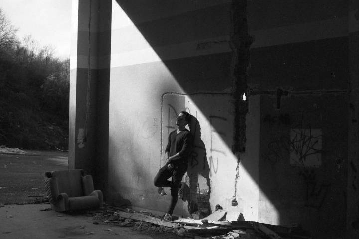 A man leans against a wall in a derelict building, illuminated by a strong diagonal beam of light.