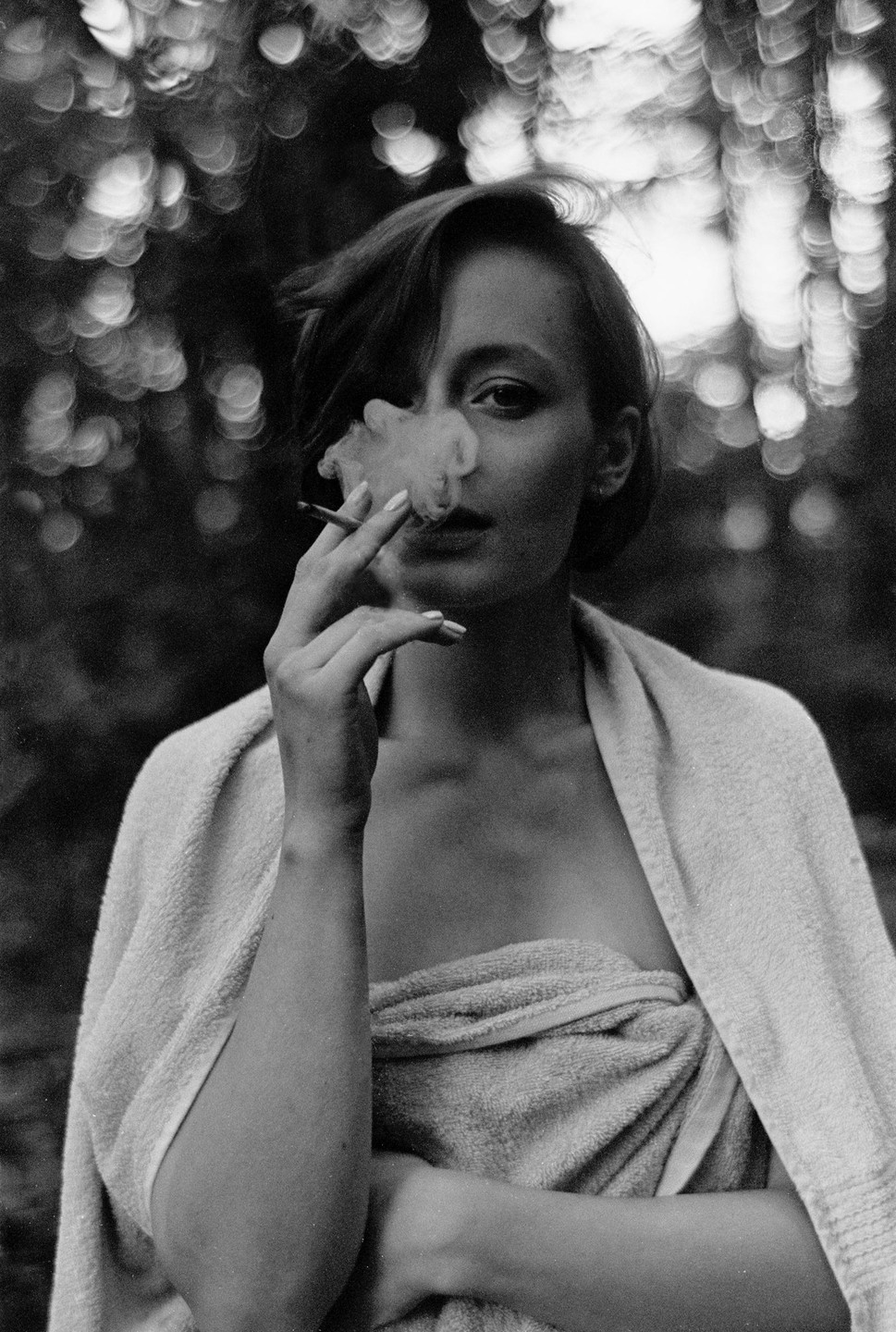 A woman wrapped in towels smokes a cigarette, with smoke obscuring part of her face, against a bokeh background.
