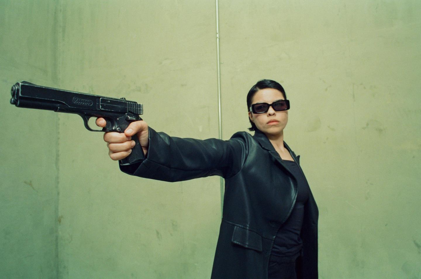 A woman in a black leather jacket and sunglasses holds a large handgun pointed forward.