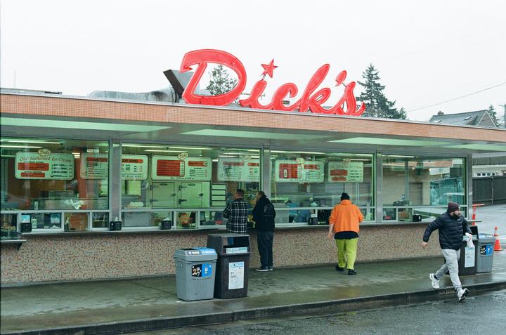 A group of men are standing outside a fast food restaurant with a red sign that says "Dick's" on a cloudy day.