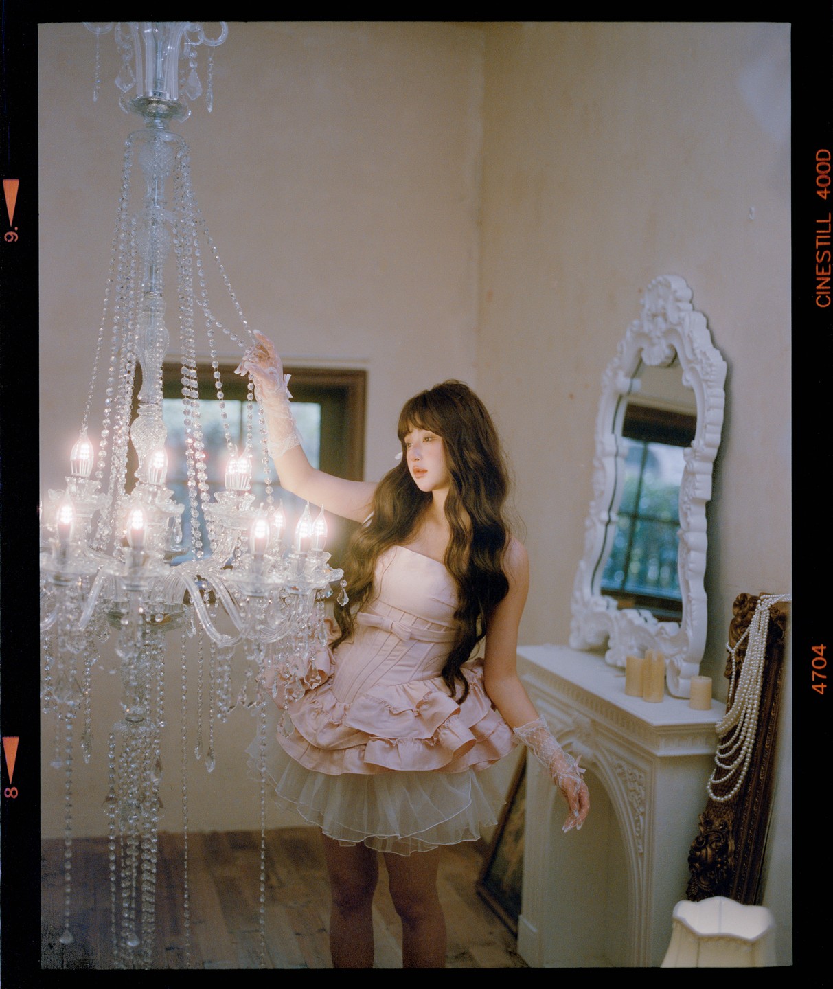 A woman in a pink dress and white gloves touches a crystal chandelier in a room with a white fireplace and mirror.