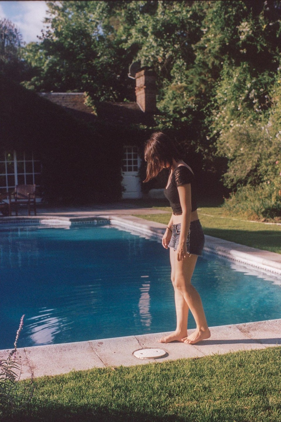 A woman stands barefoot at the edge of a swimming pool, looking down at the water, with a house and lush trees in the background.