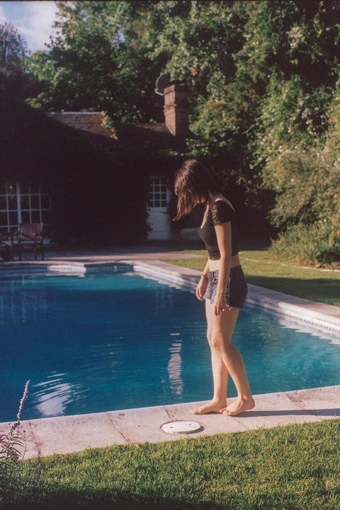 A woman stands barefoot at the edge of a swimming pool, looking down at the water, with a house and lush trees in the background.