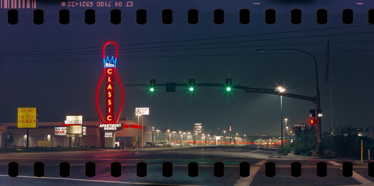 A neon bowling pin sign stands out against the night sky at an intersection with traffic lights and streetlights.
