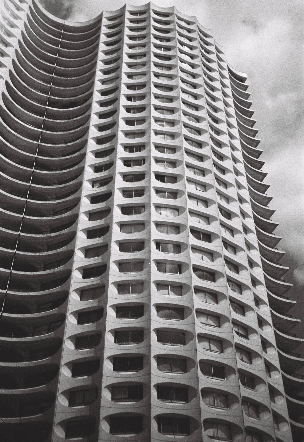 A tall, curved building with many oval windows is shown from a low angle against a cloudy sky.