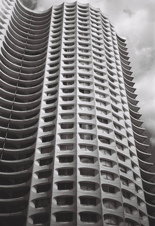 A tall, curved building with many oval windows is shown from a low angle against a cloudy sky.