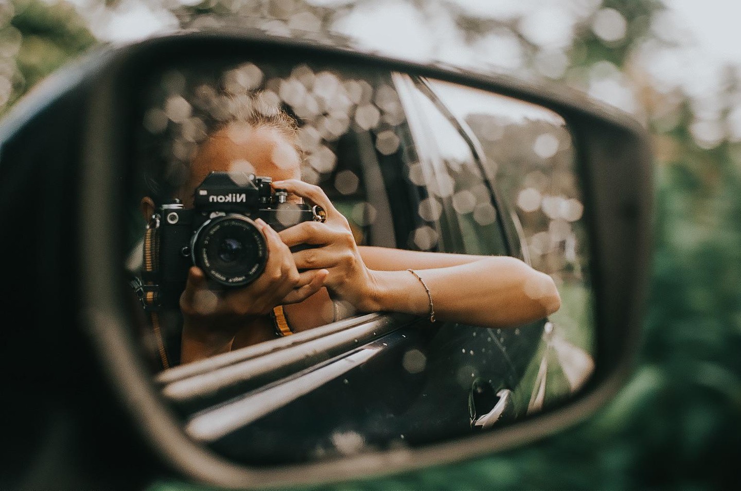 A woman holds a camera, reflected in a car's side mirror.