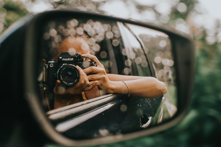 A woman holds a camera, reflected in a car's side mirror.