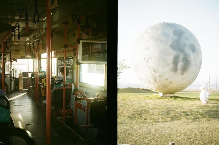 A split image shows the interior of a bus on the left and a large moon sculpture in a grassy field on the right.