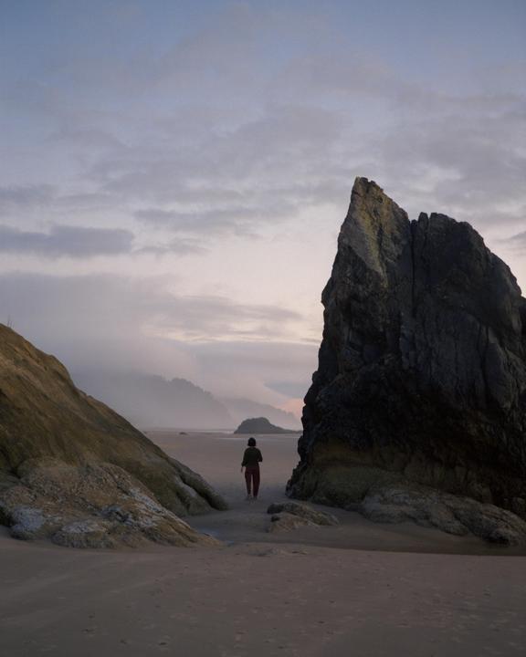 A person walks on a sandy beach between two large rock formations with a hazy mountain range in the distance.